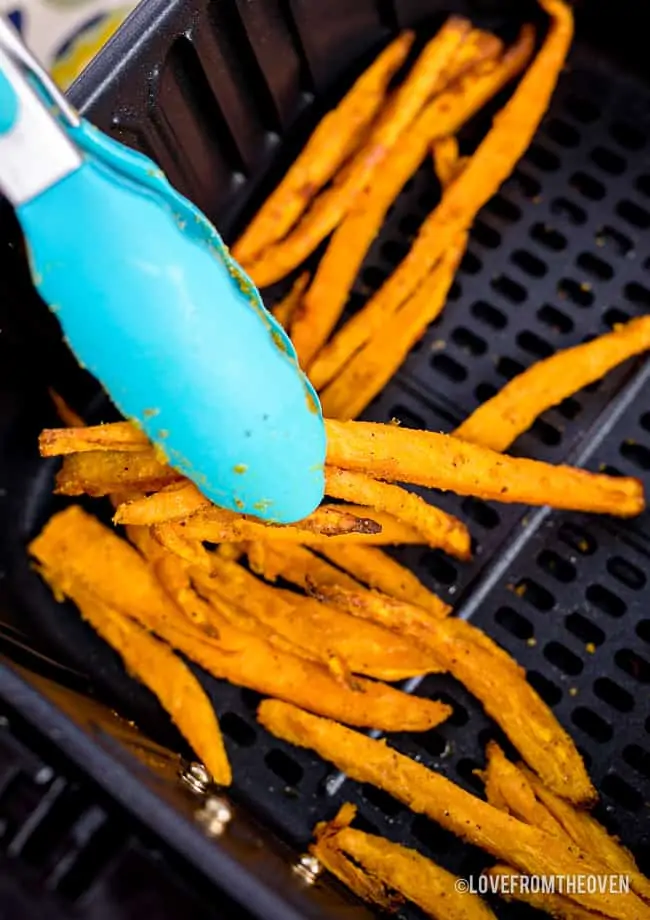 Tongs picking up sweet potato fries in an air fryer.