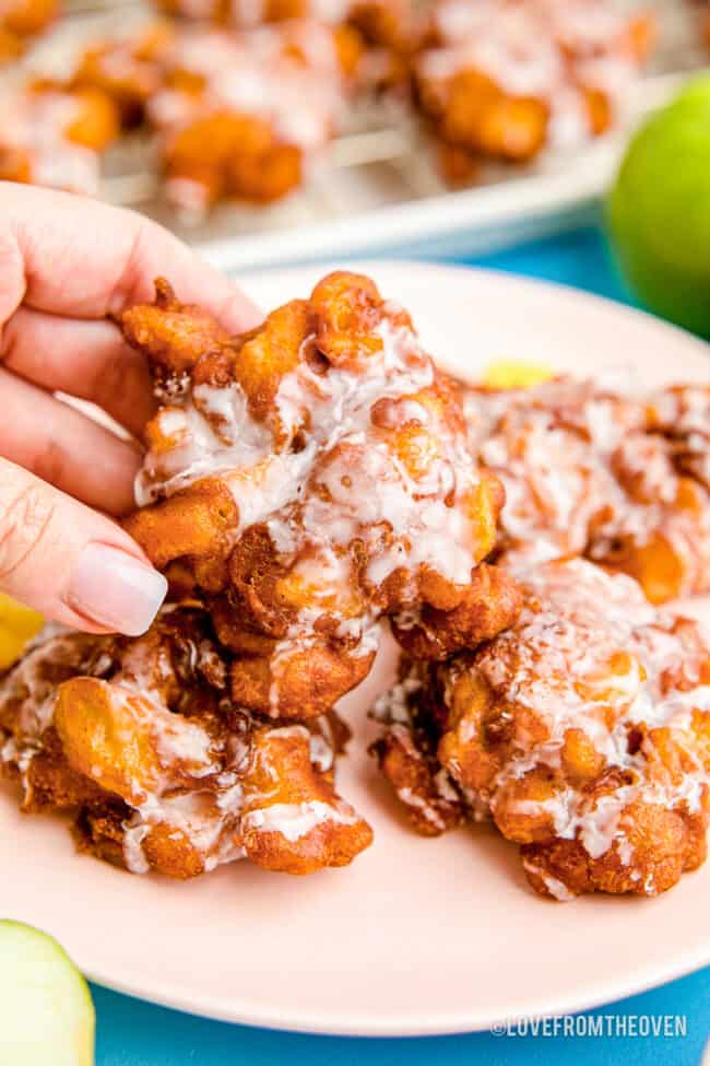 A hand grabbing an apple fritter from a plate.