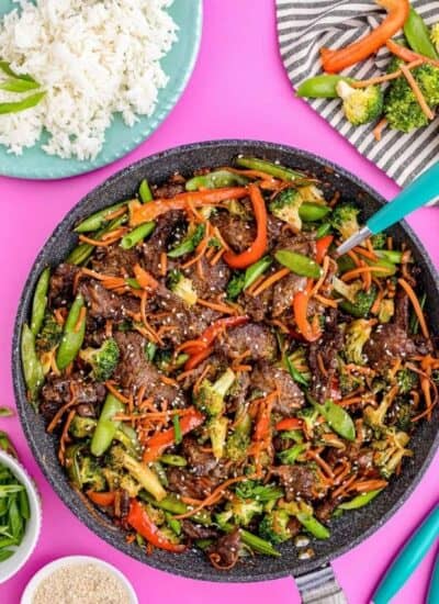 flatlay photo of a colorful stir fry dish made of beef, carrots, brocolli, capsicums, sugar snap peas and sesame seeds in a pan, set against a pink background surrounded by white rice, stir fried vegetables and condiments