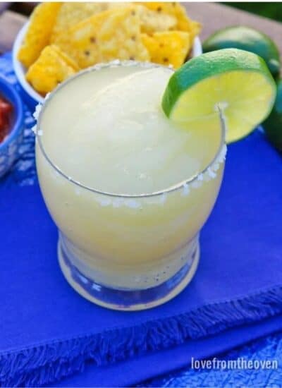 A glass of frozen margarita with a salted rim and a slice of lime as garnish, laid on a royal blue table cloth.