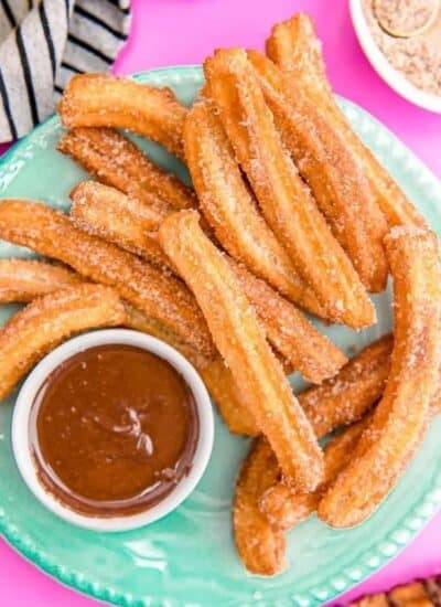 A mint green plate of golden brown churros rolled in sugar and served with a chocolate dip on the side, set in a pink background with a white tea towel on its side