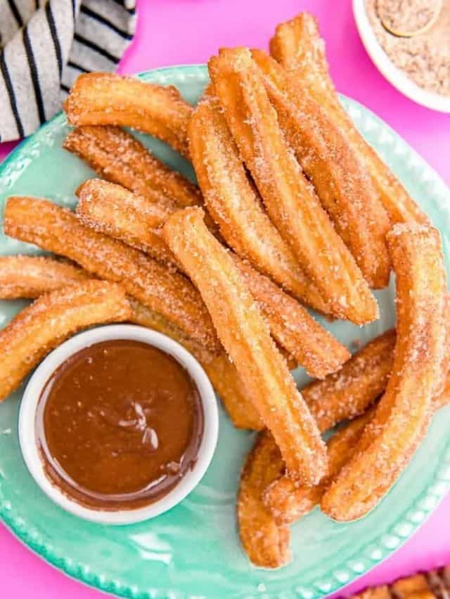 A mint green plate of golden brown churros rolled in sugar and served with a chocolate dip on the side, set in a pink background with a white tea towel on its side