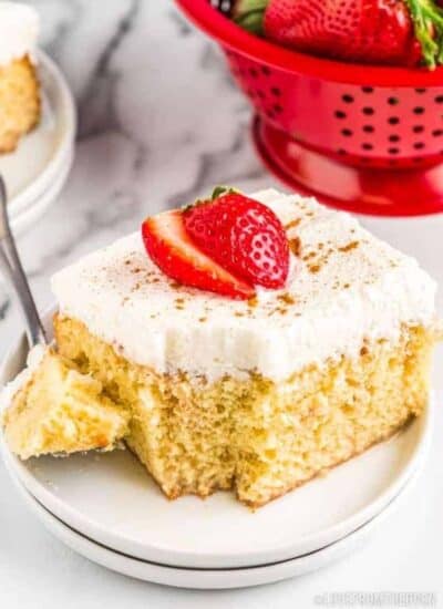 A slice of Tres Leches cake topped with a strawberry and scooped with a fork set on a marble countertop beside a red sieve full of strawberries