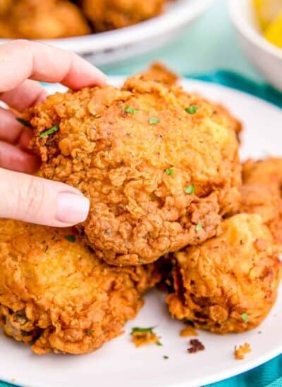 Stacks of golden brown Buttermilk Fried Chicken with one being held by a manicured hand.