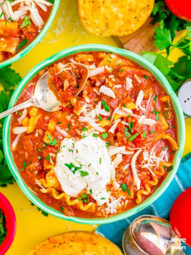 A flat lay photo of a serving of lasagna soup in a mint green bowl with a spoon dipped in it. It's surrounded with another bowl of lasagna soup, parsley, tea towels, salt and pepper shakers.