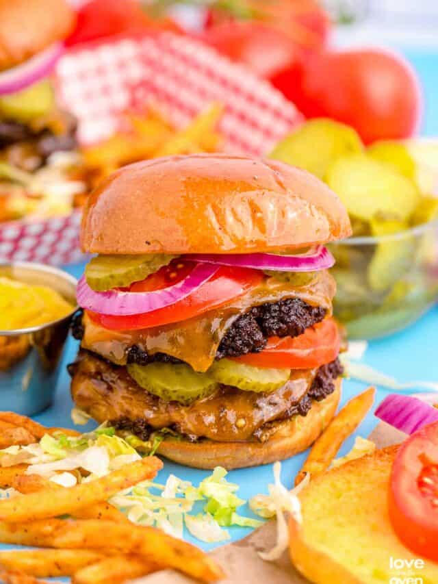 A smashburger surrounded by fries, checkered napkin and a bowl of pickles