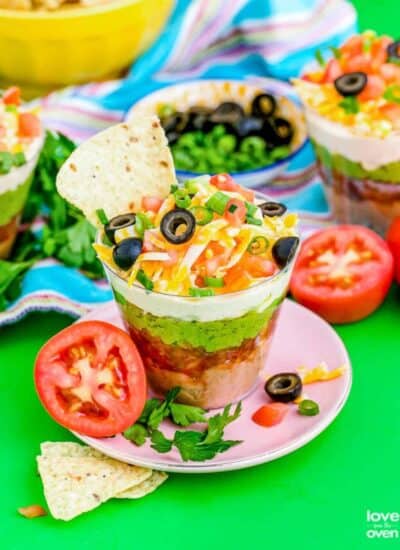 seven layer dip with a tortilla chip on a pink plate on a colorful table with tomatoes