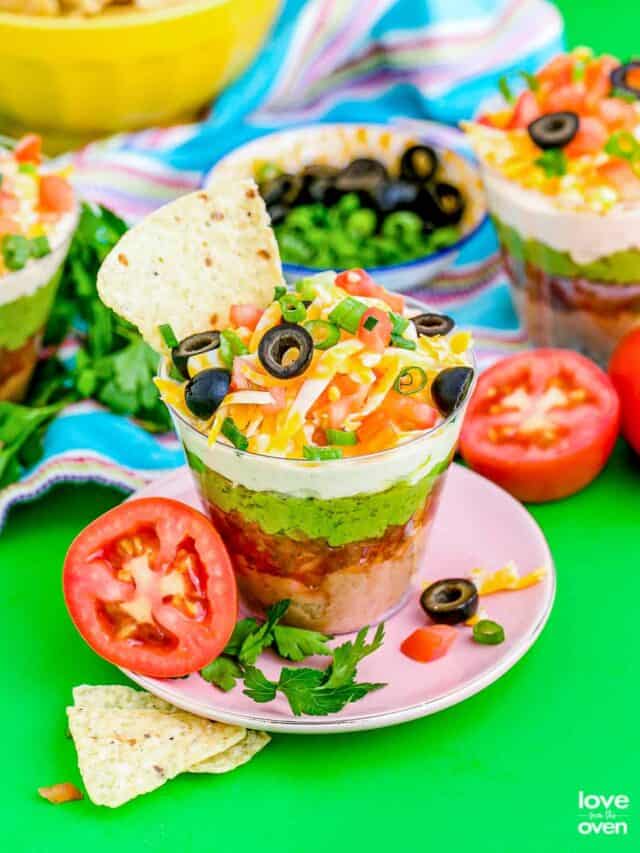 seven layer dip with a tortilla chip on a pink plate on a colorful table with tomatoes