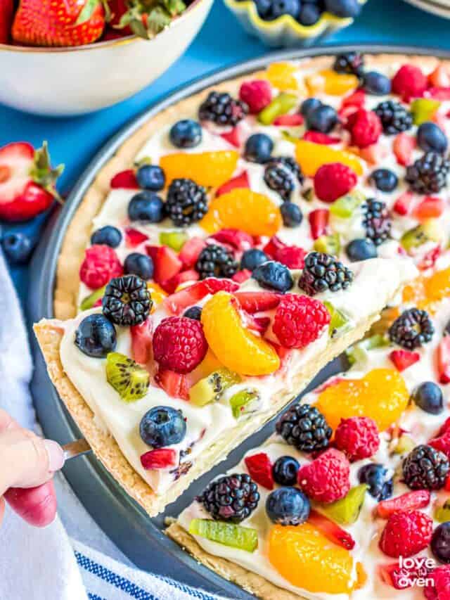 A slice of sugar cookie fruit pizza being lifted out of a pan.