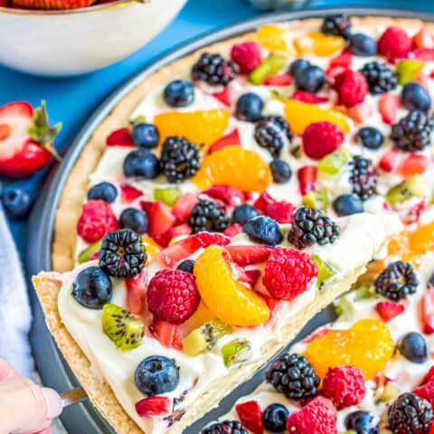 A slice of sugar cookie fruit pizza being lifted out of a pan.