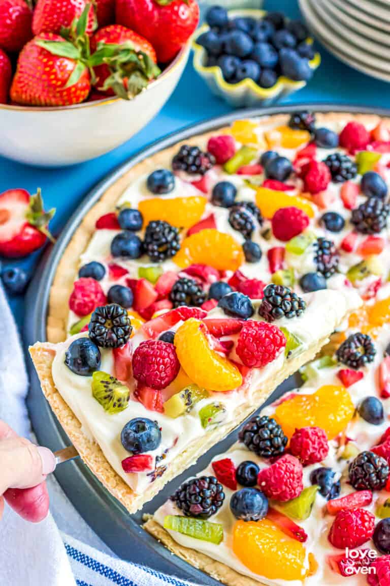 A slice of sugar cookie fruit pizza being lifted out of a pan.