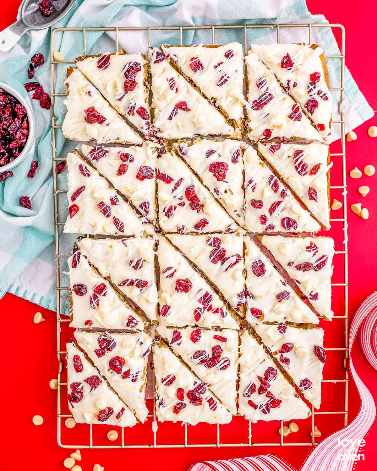 An overhead photo of a batch of cranberry bliss bars, cut and placed on a wire cooling rack, on a red background.