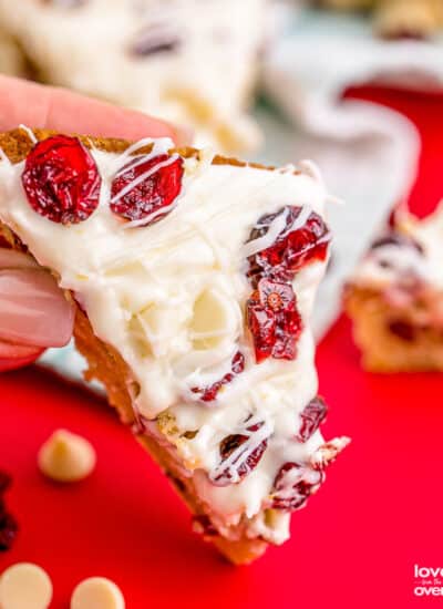 A hand holding a cranberry bliss bar on a red background.