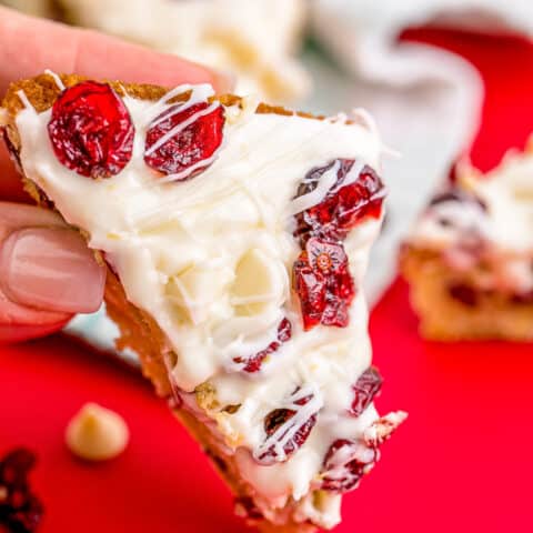 A hand holding a cranberry bliss bar on a red background.