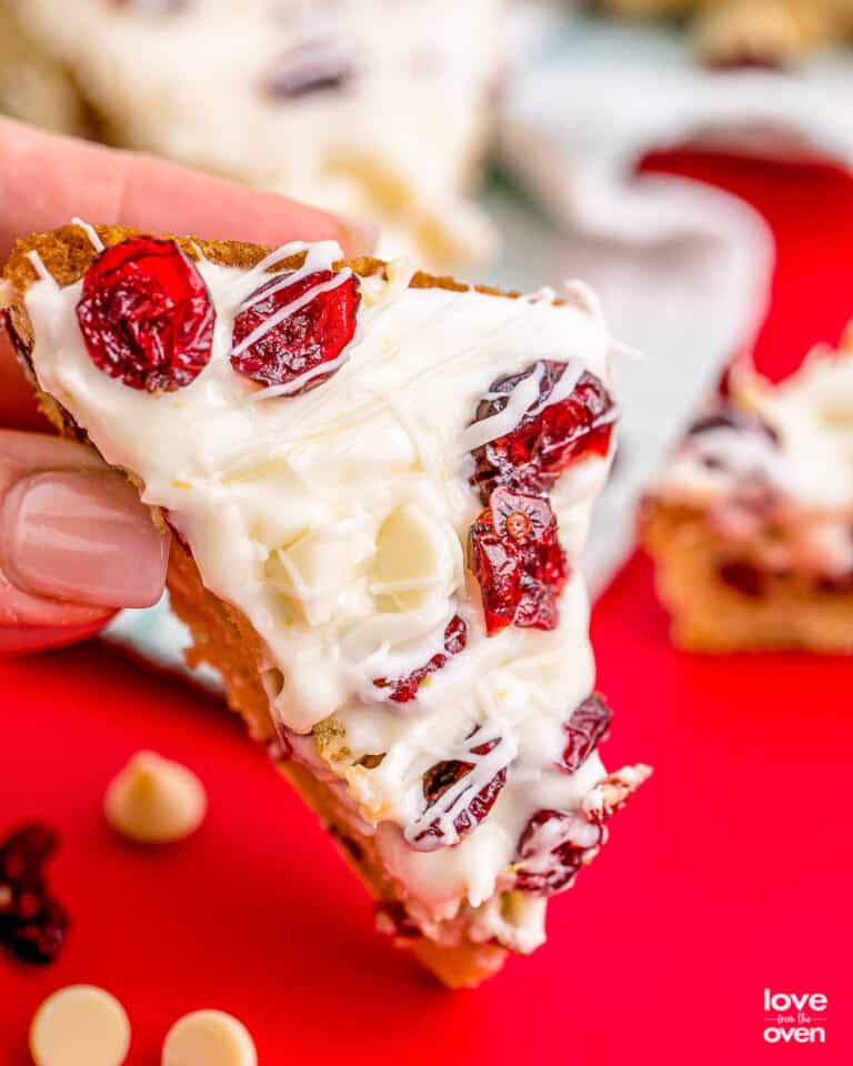 A hand holding a cranberry bliss bar on a red background.