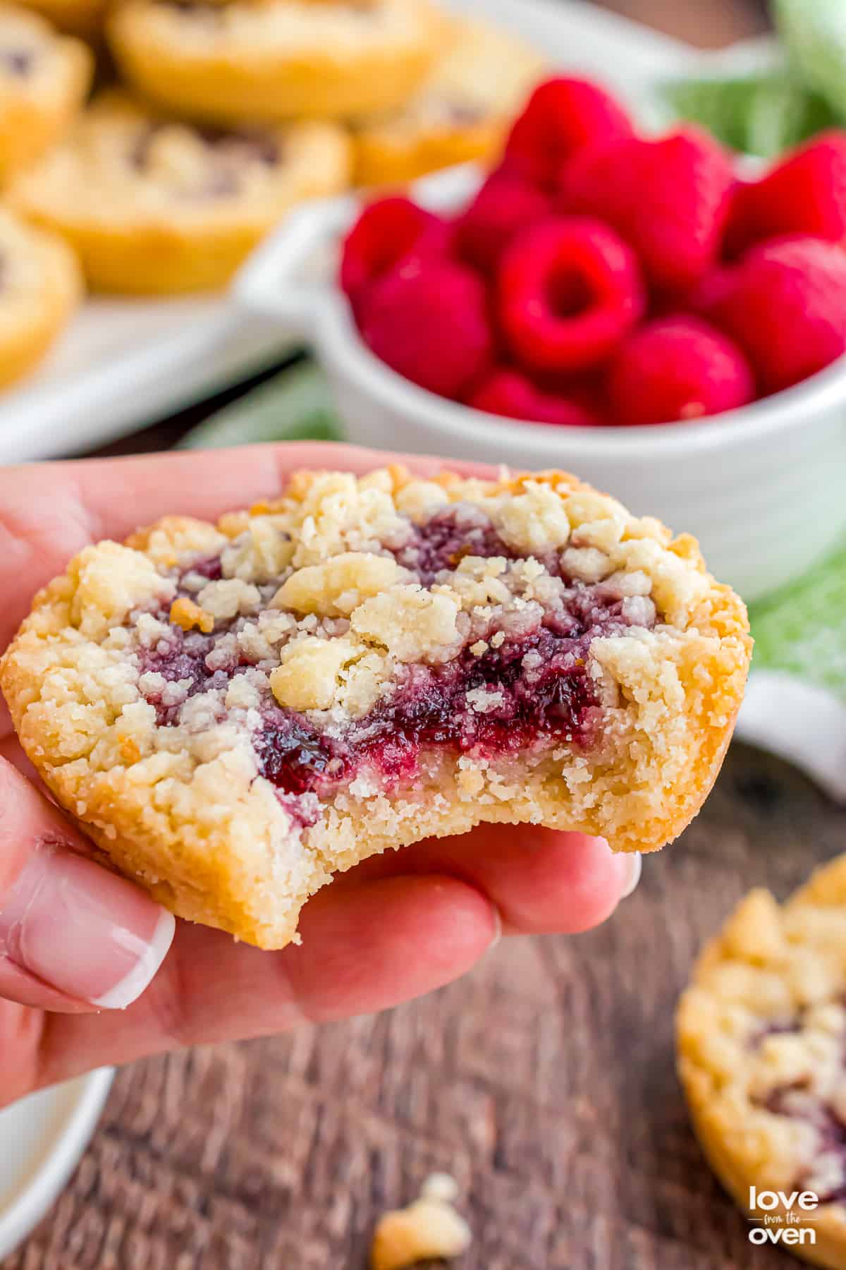 A hand holding a raspberry crumble cookie with a bite taken out of it.