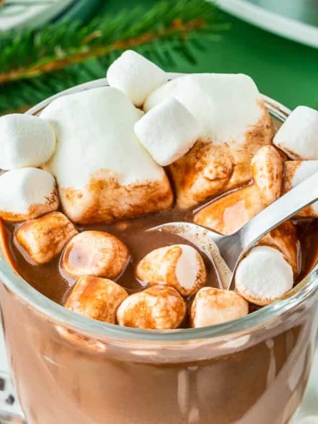 a glass of hot cocoa with marshmallows on top being stirred with a spoon