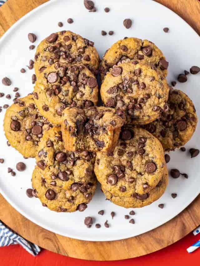 a plate of stacked banana chocolate chip muffins and the top muffin has a bite taken out of it