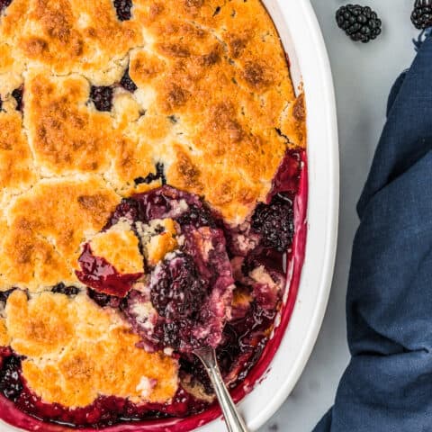 A spoon taking a serving of blackberry cobbler out of a baking dish.
