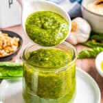 A spoon with a spoonful of homemade pesto sauce over a jar of pesto.