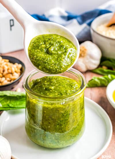 A spoon with a spoonful of homemade pesto sauce over a jar of pesto.