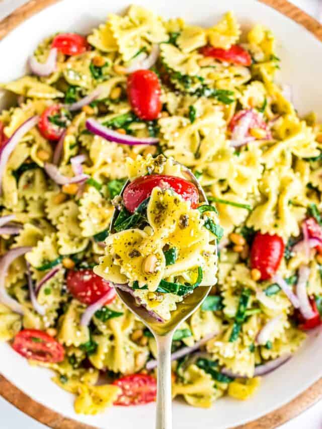 A spoon holding a serving of pesto pasta salad over a bowl full of pasta salad.