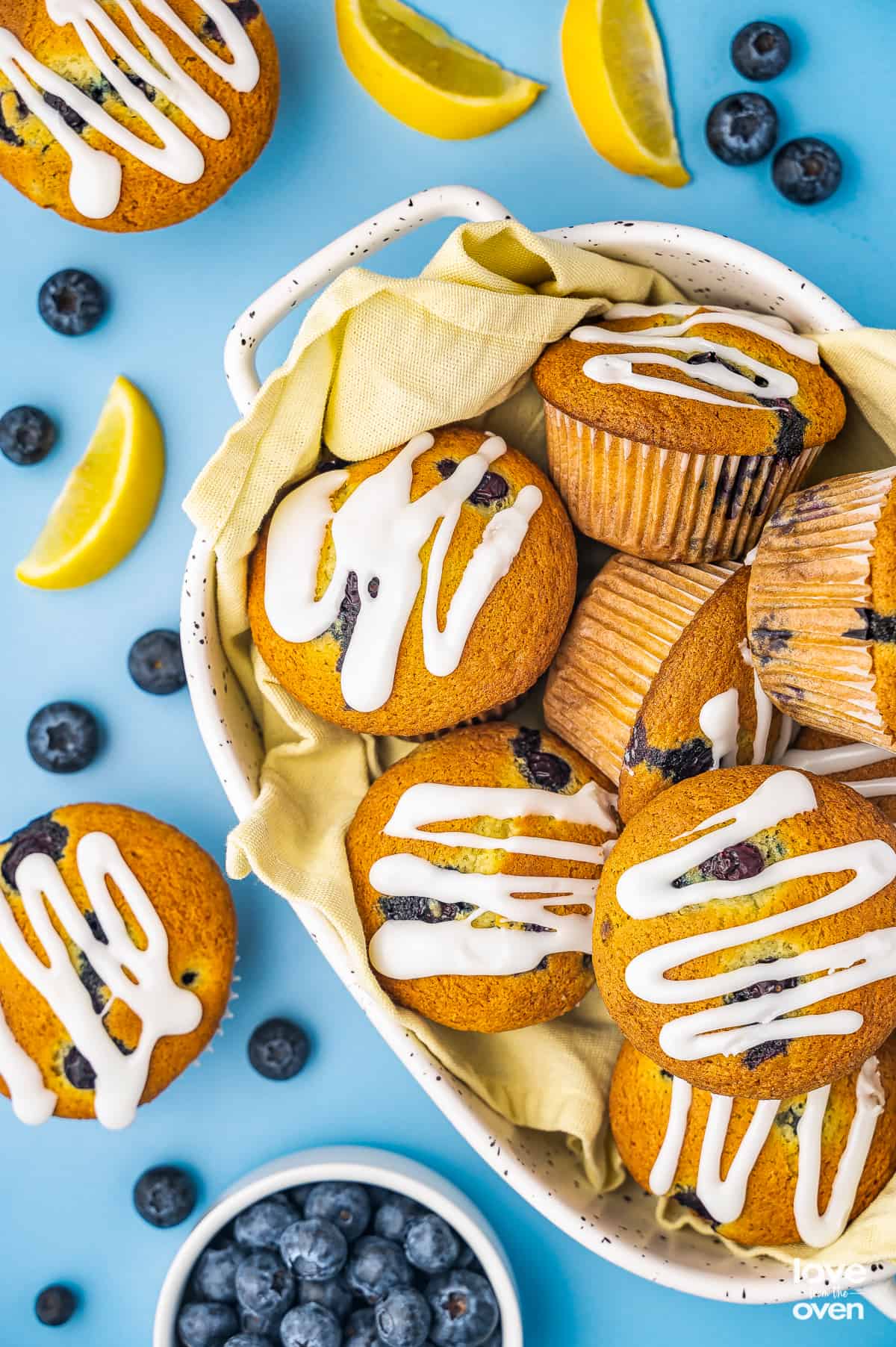 A bowl filled with lemon blueberry muffins with blueberries and lemon slices around it.