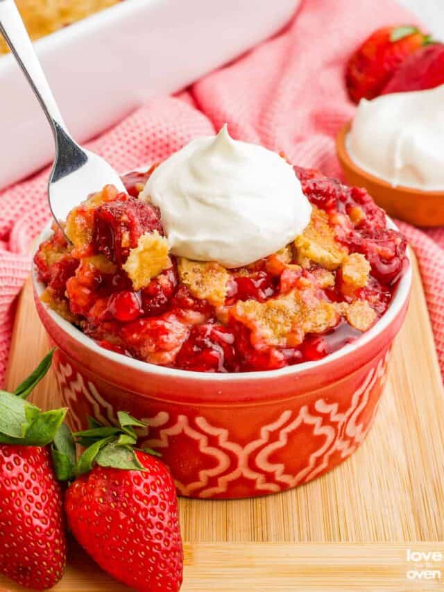 A spoon grabbing a portion of a strawberry dump cake topped with whipped cream.
