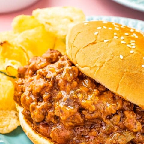 A sloppy joe on a sesame seed bun with a side of potato chips.